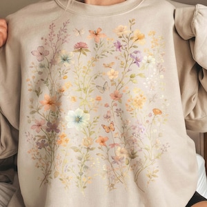 May include: A beige sweatshirt featuring a watercolor floral design with various flowers and butterflies in shades of pink, orange, yellow, and purple. The long-sleeved top has a crew neck and a relaxed fit.