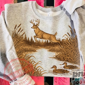 Peut inclure: Un sweat-shirt gris avec un cerf brun sautant au-dessus d'une rivi&egrave;re, avec des quenouilles et des canards dans l'eau. Le design a un aspect vintage et plein air. Le sweat-shirt est fait d'une mati&egrave;re douce.