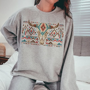 May include: A grey sweatshirt with a Southwestern design. It features a bull skull with turquoise, red, and brown geometric patterns. The sweatshirt has long sleeves and a crew neck. The design is reminiscent of Native American art.