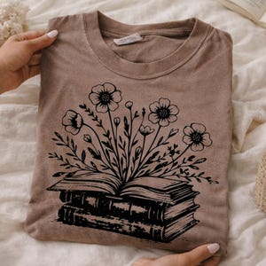 Pode incluir: Uma camiseta marrom com um design floral preto emergindo de um livro aberto. O gráfico apresenta flores e folhas detalhadas, com o livro aberto sobre uma pilha de livros. A camiseta é feita de material macio.