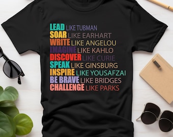 Może przedstawiać: Czarny T-shirt z kolorowym wzorem graficznym z tekstem "Lead like Tubman, Soar like Earhart, Write like Angelou, Imagine like Kahlo, Discover like Curie, Speak like Ginsburg, Inspire like Yousafzai, Be Brave like Bridges, Challenge like Parks".