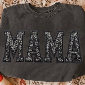 May include: Dark gray sweatshirt with the word "MAMA" in large, glittery, silver letters. The letters are outlined in a darker gray, creating a bold contrast. The sweatshirt has a crew neck and a relaxed fit.