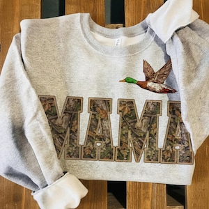 May include: A gray sweatshirt with the word "MAMA" in camouflage letters. A flying duck with green, brown, and red details is above the text. The sweatshirt has a crew neck and white cuffs.