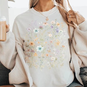 May include: A cream-colored sweatshirt featuring a watercolor floral design with pastel-colored flowers and butterflies. The flowers include shades of blue, yellow, purple, and white. The sweatshirt has a crew neck and long sleeves.