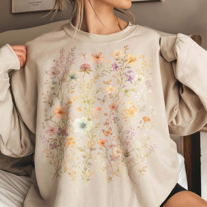 May include: A beige sweatshirt featuring a watercolor floral design with various flowers and butterflies in shades of pink, orange, yellow, and purple. The long-sleeved top has a crew neck and a relaxed fit.