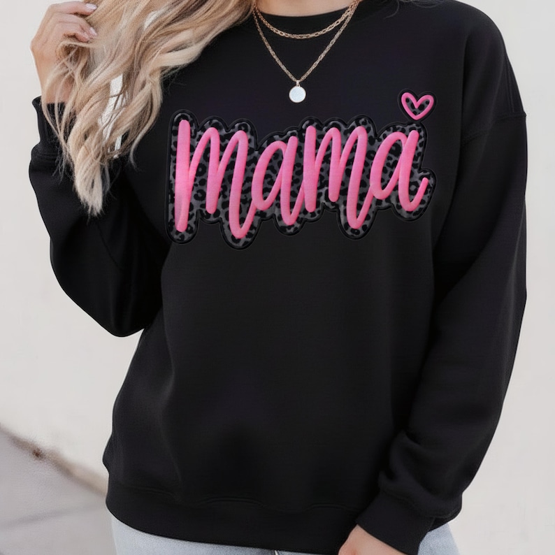 Op de afbeelding: Zwarte sweatshirt met het woord "Mama" in roze script met een luipaardprint. Een klein roze hartje staat boven de letter "a". De sweatshirt heeft lange mouwen en een ronde hals.