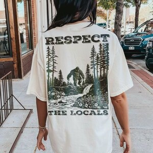 May include: Cream-colored t-shirt with a Bigfoot graphic and the text "RESPECT THE LOCALS." The design features a Bigfoot standing near a waterfall and trees. The shirt has short sleeves and a relaxed fit.