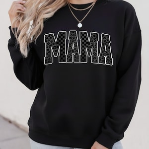 May include: Black sweatshirt with the word "MAMA" in large, bold letters across the front. The letters have a checkered and leopard print design with a white outline. The sweatshirt is a casual style.