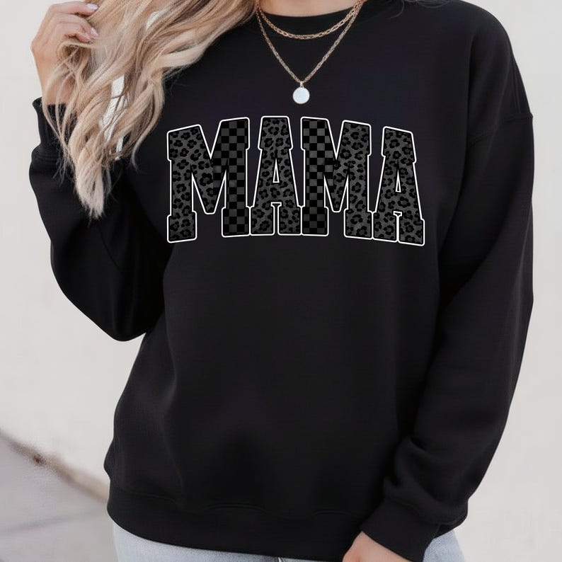 May include: Black sweatshirt with the word "MAMA" in large, bold letters across the front. The letters have a checkered and leopard print design with a white outline. The sweatshirt is a casual style.
