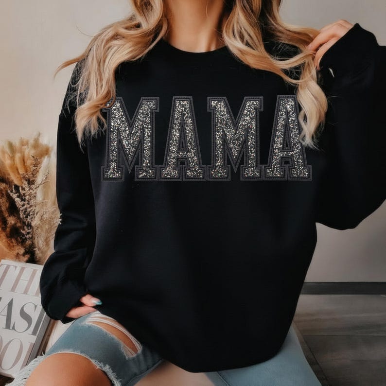 May include: Black sweatshirt with the word "MAMA" in large, glittery silver letters across the chest. The letters have a dark outline, creating a bold contrast. The sweatshirt is a casual, comfortable style.