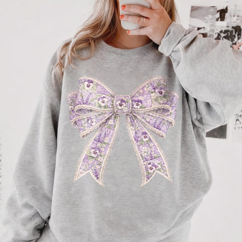May include: A gray sweatshirt featuring a large, detailed bow design. The bow is primarily lavender with floral and lace details. The design is centered on the front of the sweatshirt, creating a visually appealing and feminine aesthetic.