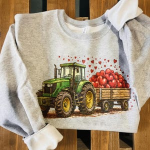 May include: A light gray sweatshirt featuring a green tractor pulling a wooden trailer filled with red heart-shaped balloons. The tractor has yellow wheels and is set against a brown background.