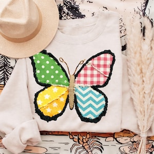 Può includere: Una felpa color crema presenta un'applicazione a farfalla grande e colorata. Le ali della farfalla sono patchwork con pois verdi, stelle gialle, motivo a quadri rossi e motivi a zig-zag blu. Un cappello beige poggia sul lato sinistro della felpa.