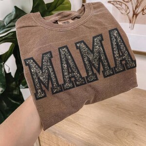 May include: A brown t-shirt with the word "MAMA" in large, glittery, black-outlined letters. The shirt has a vintage, slightly faded appearance. The t-shirt is made of a soft material.
