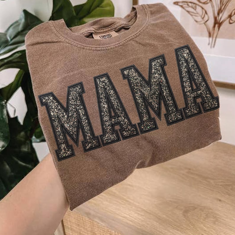 May include: A brown t-shirt with the word "MAMA" in large, glittery, black-outlined letters. The shirt has a vintage, slightly faded appearance. The t-shirt is made of a soft material.