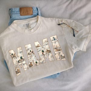 May include: Cream-colored t-shirt with the word "MAMA" in large, block letters. Each letter is filled with a detailed illustration of ducks in a snowy landscape. The shirt is paired with light blue jeans and round-framed glasses.