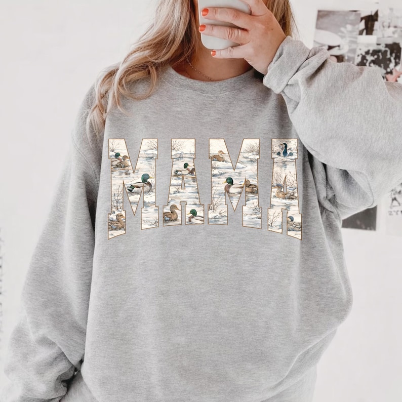 May include: A light gray sweatshirt with the word "MAMA" printed across the chest. The letters are filled with a duck-themed illustration. The sweatshirt has long sleeves and a crew neck.