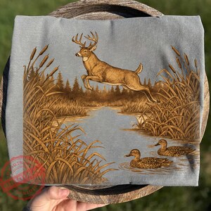 Peut inclure: T-shirt bleu clair avec une illustration d&eacute;taill&eacute;e d'un cerf sautant au-dessus d'une rivi&egrave;re, avec des canards nageant dans l'eau. Le motif comprend des quenouilles et une for&ecirc;t en arri&egrave;re-plan, cr&eacute;ant une sc&egrave;ne sur le th&egrave;me de la nature.