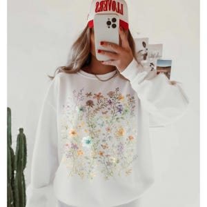 May include: White sweatshirt featuring a colorful floral design with butterflies. The design includes various flowers in shades of yellow, pink, purple, and blue. The sweatshirt has long sleeves and a crew neck.
