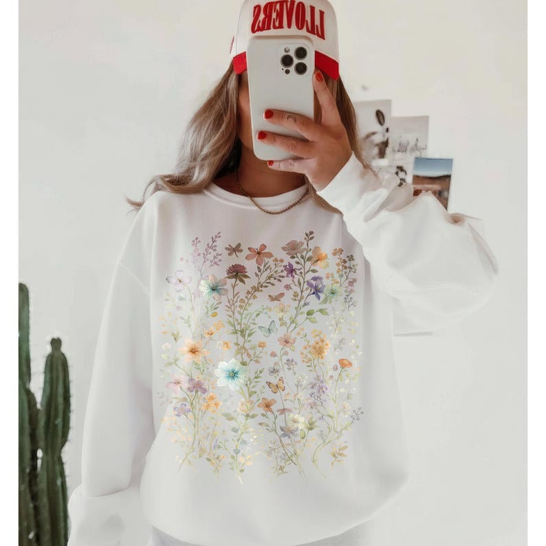 May include: White sweatshirt featuring a colorful floral design with butterflies. The design includes various flowers in shades of yellow, pink, purple, and blue. The sweatshirt has long sleeves and a crew neck.