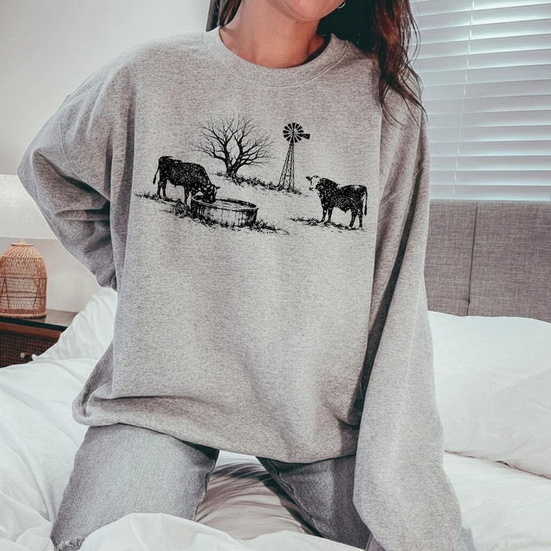 May include: A light gray sweatshirt featuring a black and white illustration of a rural scene. The design includes two cows, a water trough, a bare tree, and a windmill. The sweatshirt is made of a soft material.