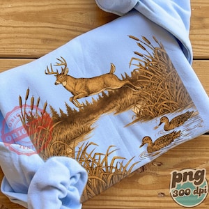 Peut inclure: Sweat-shirt bleu clair avec une illustration d&eacute;taill&eacute;e d'un cerf sautant au-dessus de l'eau. Le motif comprend des canards, des roseaux et une sc&egrave;ne de for&ecirc;t, dans des tons bruns et beiges.