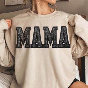 May include: Beige sweatshirt with the word "MAMA" in large, glittery, black block letters across the chest. The sweatshirt has long sleeves and a crew neck. The overall style is casual and comfortable.