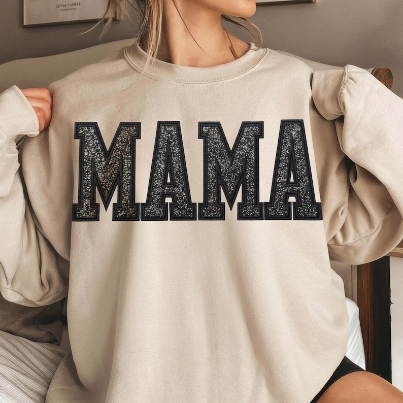 May include: Beige sweatshirt with the word "MAMA" in large, glittery, black block letters across the chest. The sweatshirt has long sleeves and a crew neck. The overall style is casual and comfortable.