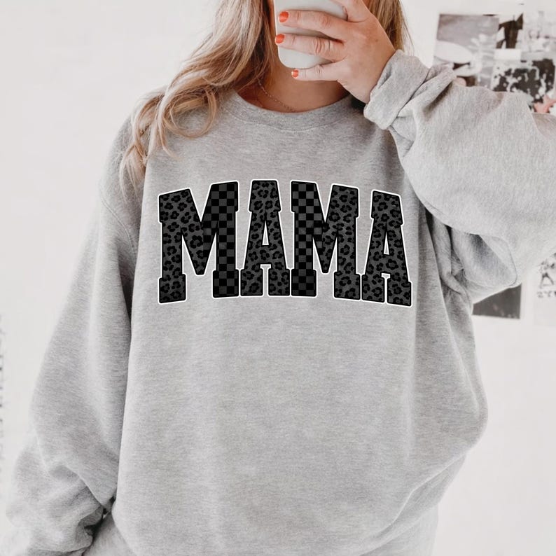 May include: A light gray sweatshirt with the word "MAMA" in large, bold letters across the front. The letters have a black and white checkered pattern and a black leopard print design. The sweatshirt has long sleeves.
