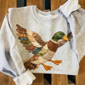 May include: A light gray sweatshirt featuring a detailed appliqué of a flying duck. The duck has a green head, brown and colorful wings, and orange feet. The sweatshirt has a crew neck and long sleeves with white cuffs.