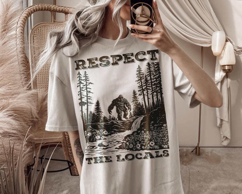May include: Off-white t-shirt with a Bigfoot graphic. The design features a forest scene with the text "RESPECT THE LOCALS" in a vintage font. The graphic is in shades of brown and black.