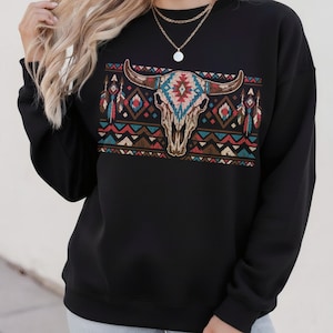 May include: Black sweatshirt with a bull skull graphic, adorned with turquoise, red, and brown tribal patterns. The design incorporates feathers and geometric shapes, evoking a Southwestern style. The sweatshirt has a crew neck and long sleeves.