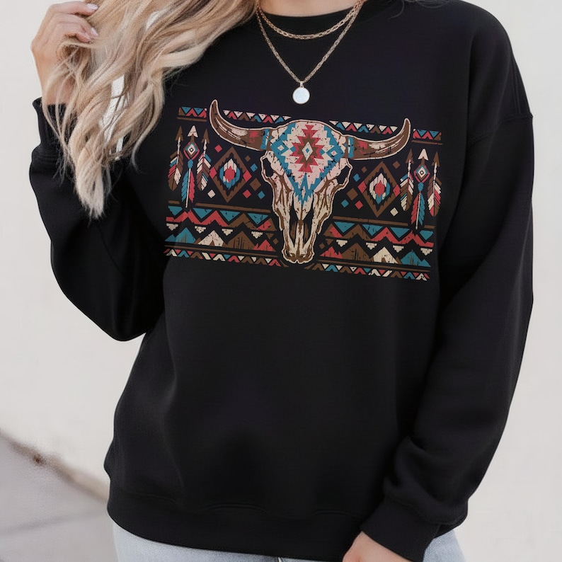 May include: Black sweatshirt with a bull skull graphic, adorned with turquoise, red, and brown tribal patterns. The design incorporates feathers and geometric shapes, evoking a Southwestern style. The sweatshirt has a crew neck and long sleeves.
