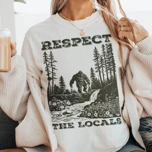May include: Cream-colored sweatshirt with a black graphic featuring a Bigfoot illustration in a forest scene. The text "RESPECT THE LOCALS" is printed above and below the image. The design is centered on the front of the shirt.