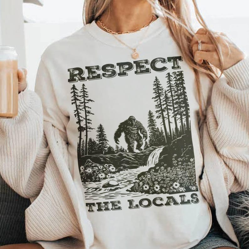May include: Cream-colored sweatshirt with a black graphic featuring a Bigfoot illustration in a forest scene. The text "RESPECT THE LOCALS" is printed above and below the image. The design is centered on the front of the shirt.