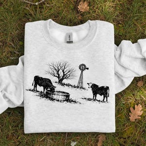 May include: A light gray sweatshirt featuring a black and white illustration of a rural scene. The design includes two cows, a tree, a windmill, and a water trough. The sweatshirt is made of a soft material.