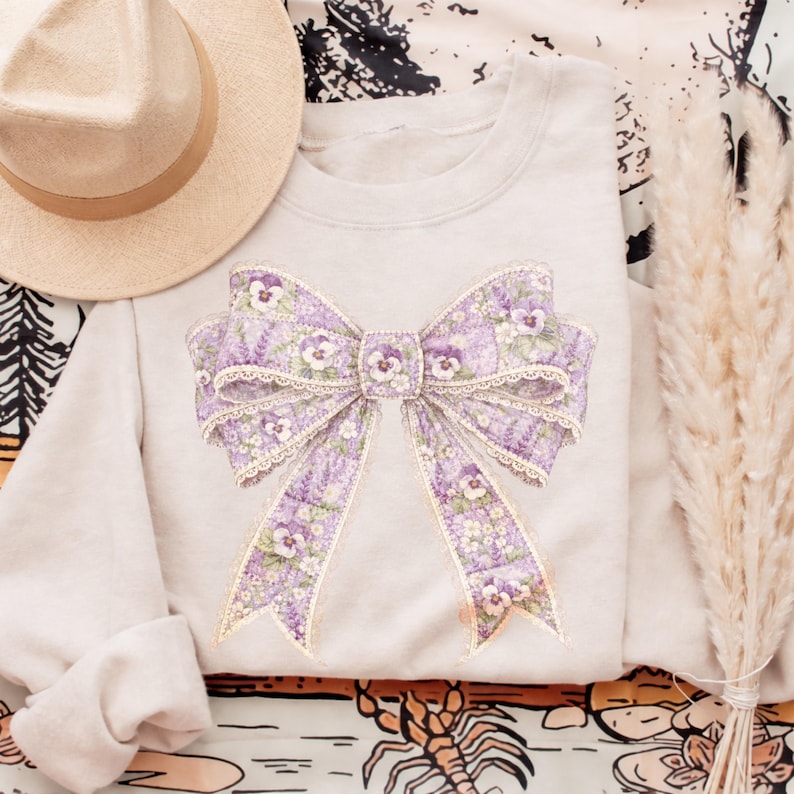 May include: A light gray sweatshirt featuring a large, detailed bow design in shades of lavender and white. The bow is adorned with floral patterns and lace trim. A beige hat rests on the top left. The sweatshirt is folded, with the sleeves tucked in.