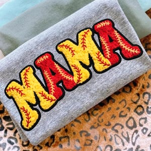 May include: Grey sweatshirt with the word "MAMA" in yellow and red baseball-themed lettering. The letters are outlined in black and feature baseball stitching details. The sweatshirt is folded, with other fabric visible in the background.