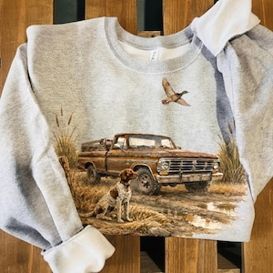 Puede incluir: Una sudadera gris con un gráfico detallado de una camioneta marrón vintage, un perro de caza y un pato volando. El diseño está ambientado en una escena natural al aire libre con hierba alta y un camino embarrado. La sudadera tiene cuello redondo y mangas largas.