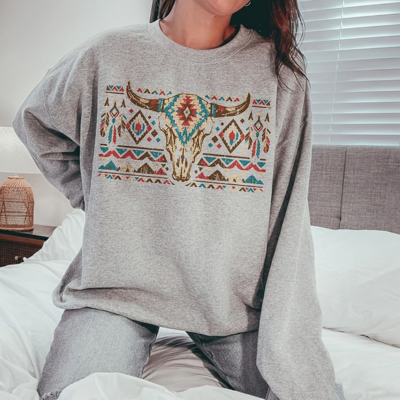 May include: A grey sweatshirt with a Southwestern design. It features a bull skull with turquoise, red, and brown geometric patterns. The sweatshirt has long sleeves and a crew neck. The design is reminiscent of Native American art.