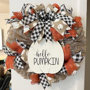 May include: A burlap wreath decorated with black and white buffalo check ribbon, orange ribbon, and white pumpkins with brown polka dots. The wreath has a white sign in the center that says "hello pumpkin".