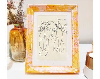 May include: A rectangular picture frame with a marbled yellow and pink pattern containing a black line drawing of a person with a floral crown on a cream background. The drawing includes the handwritten text 15 mai 58 in the upper right corner.