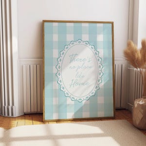 Puede incluir: Un estampado enmarcado con un fondo de patrón de cuadros vichy azul claro y blanco. El estampado tiene un diseño ovalado con las palabras "There's no place like Home" en una fuente de escritura. El marco es de color marrón cálido.