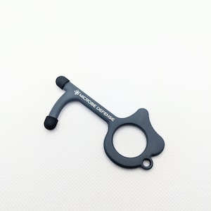 Pode incluir: Uma ferramenta cinza para abrir portas com uma ponta de borracha preta em cada extremidade. A ferramenta tem um laço em uma extremidade e o texto "MICROBE DEFENSE" impresso no lado.