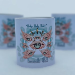 Pode incluir: Caneca de cerâmica branca com uma ilustração detalhada de múltiplos olhos com asas emplumadas, rodeados por um desenho circular turquesa e branco. O texto "Holy Holy Holy" e "Isaías 63" estão impressos na caneca.