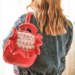May include: A coral-colored crochet backpack with a woven, light pink and white center panel. The bag has a ruffled edge and a handle. The person is wearing a denim jacket. The bag is a unique, handmade accessory.