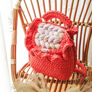 May include: Coral-colored crochet handbag with a woven center panel of white and pink. The bag has a ruffled edge and a matching handle. It is sitting on a wicker chair.