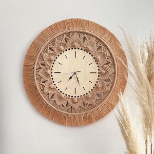 Puede incluir: Un reloj de pared de macramé beige con una cara de madera y manecillas negras. El reloj está rodeado por una franja de hilo beige.