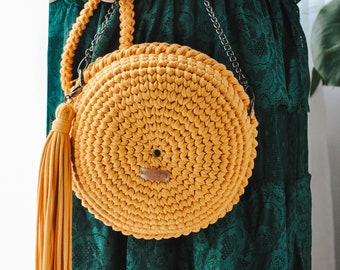 May include: A round, crocheted handbag with a mustard yellow color and a long tassel. The bag has a silver chain strap.