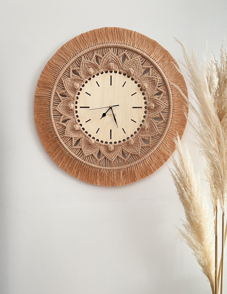 May include: A beige macrame wall clock with a wooden face and black hands. The clock is surrounded by a fringe of beige yarn.